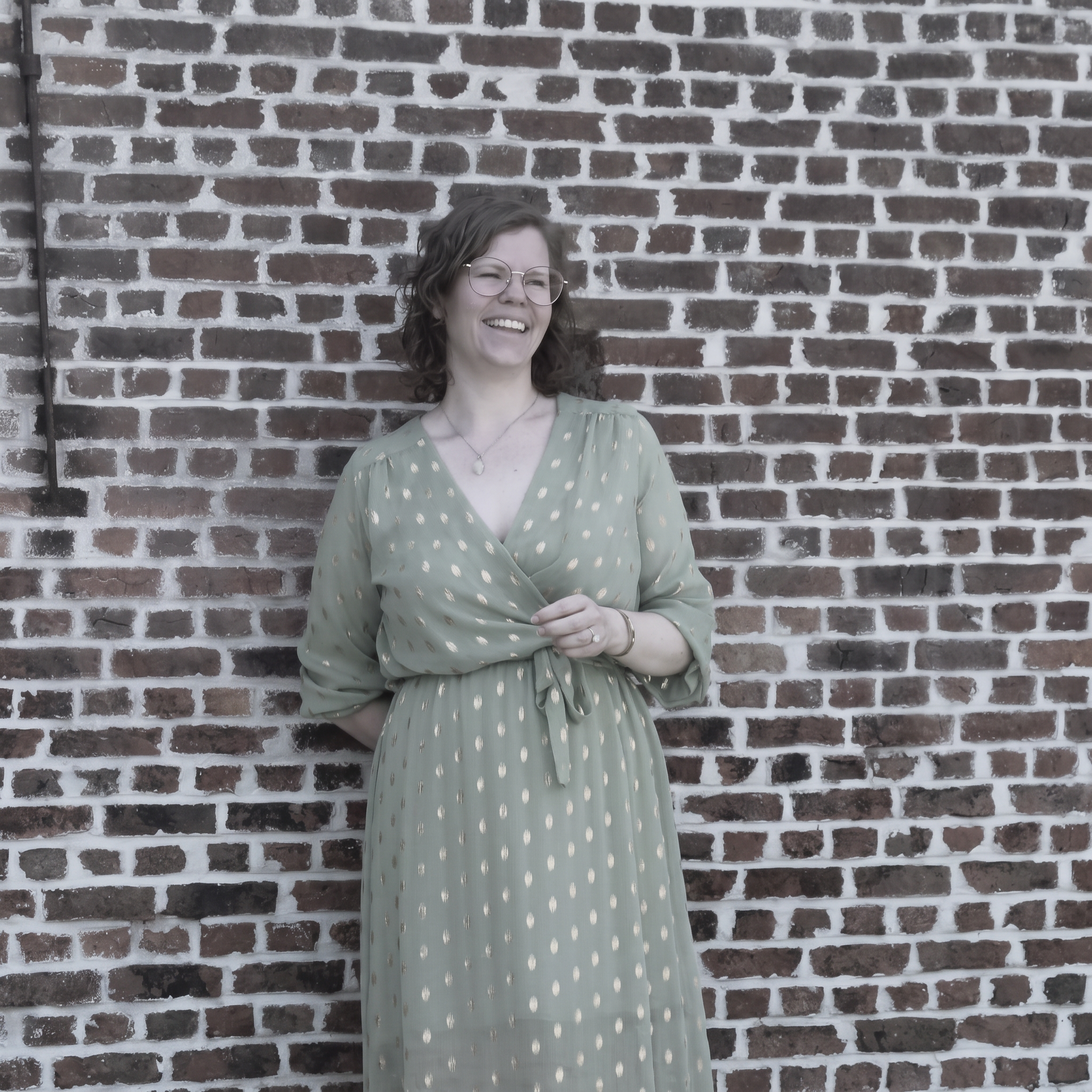 Maike lacht voor een bakstenen muur in een saliegroene jurk