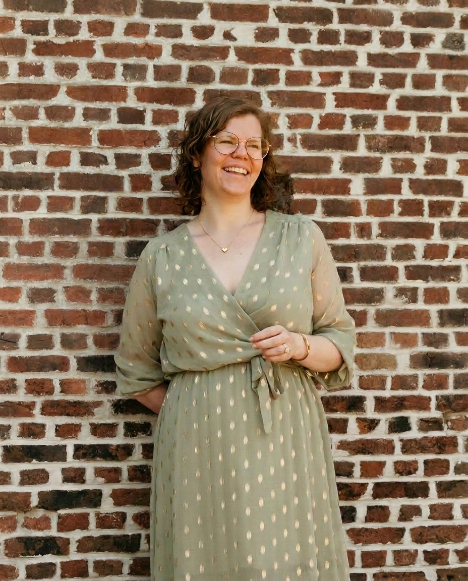 Maike - warm lachend in een saliegroene jurk tegen een geschilderde bakstenen muur
